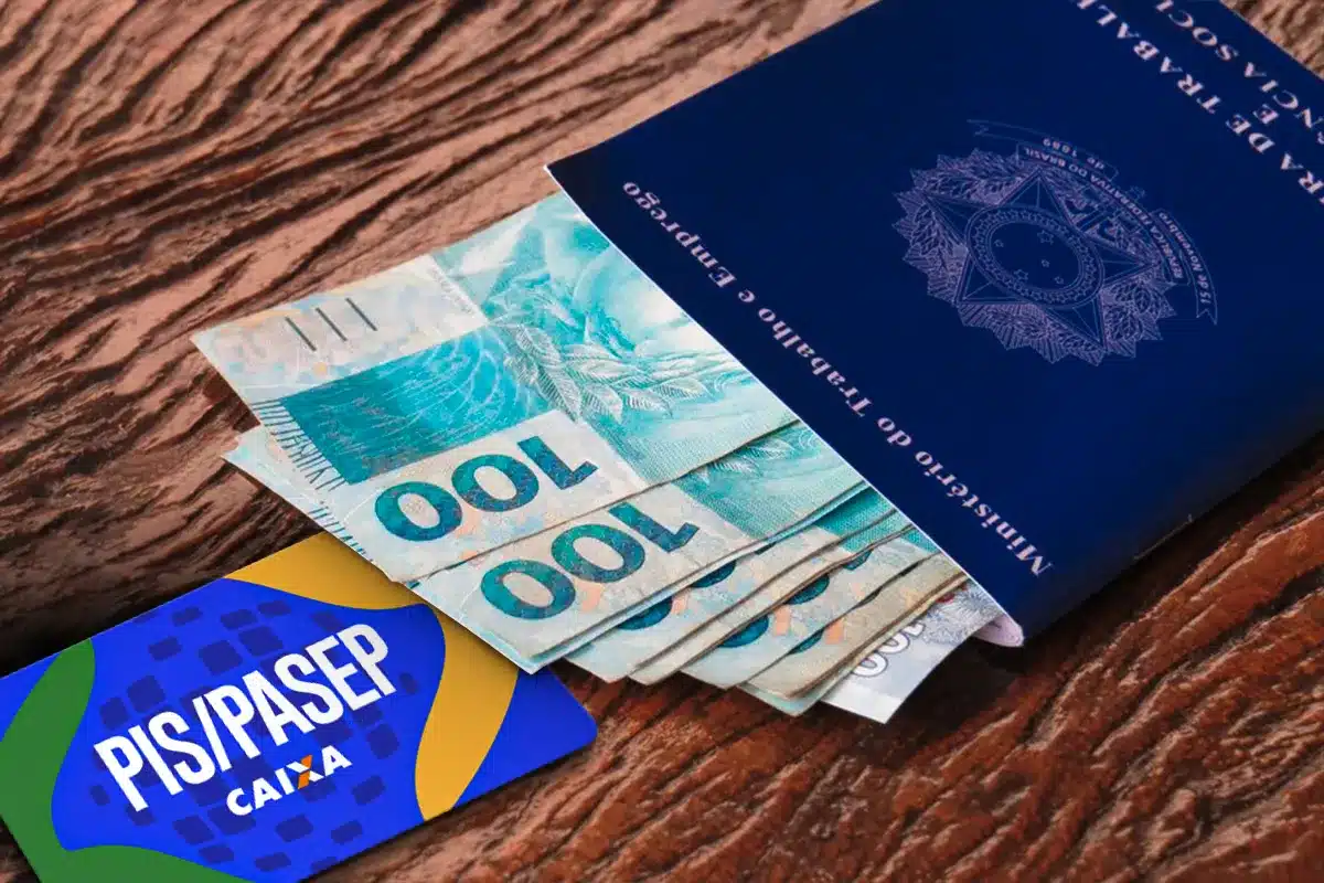 PIS/PASEP com carteira de trabalho e cédulas de dinheiro Carteira de trabalho azul, dinheiro em cédulas de cem reais e cartão PIS/PASEP sobre mesa de madeira