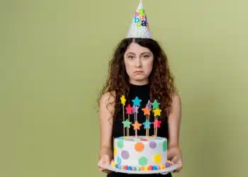 Pessoa com expressão de desânimo segurando um bolo de aniversário decorado com estrelas coloridas e bolinhas de massa, usando um chapéu de festa.