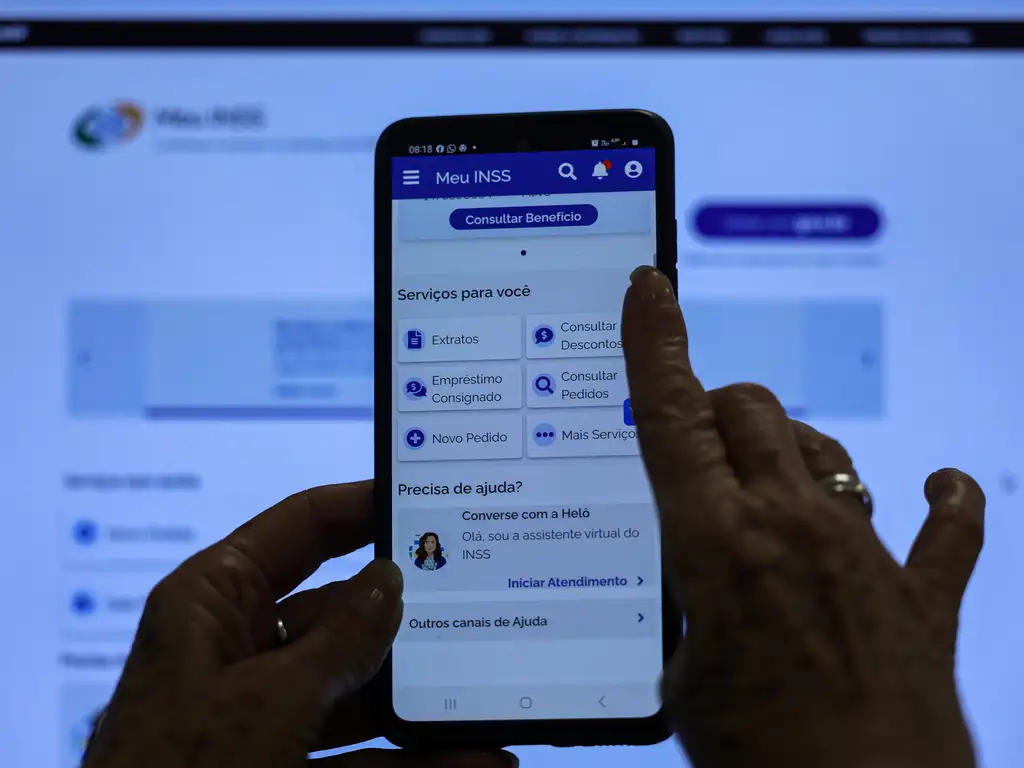 Mãos segurando um celular com o aplicativo oficial "Meu INSS" aberto na tela principal de serviços e consulta de benefícios.