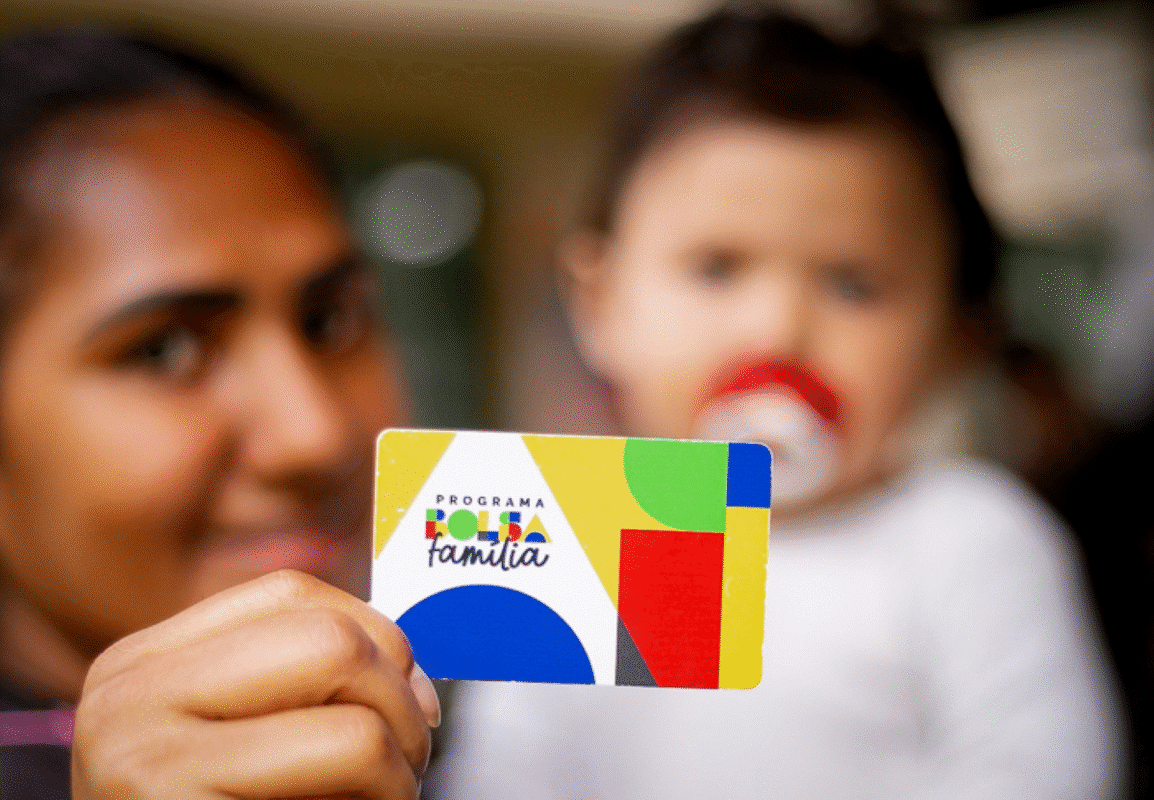 Mulher segura o cartão do Programa Bolsa Família com um bebê ao fundo.