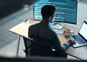 Desenvolvedor ou analista de sistemas trabalhando à noite em frente a um computador, com a tela principal exibindo linhas de código, simbolizando o foco em tecnologia e futuro do trabalho digital.