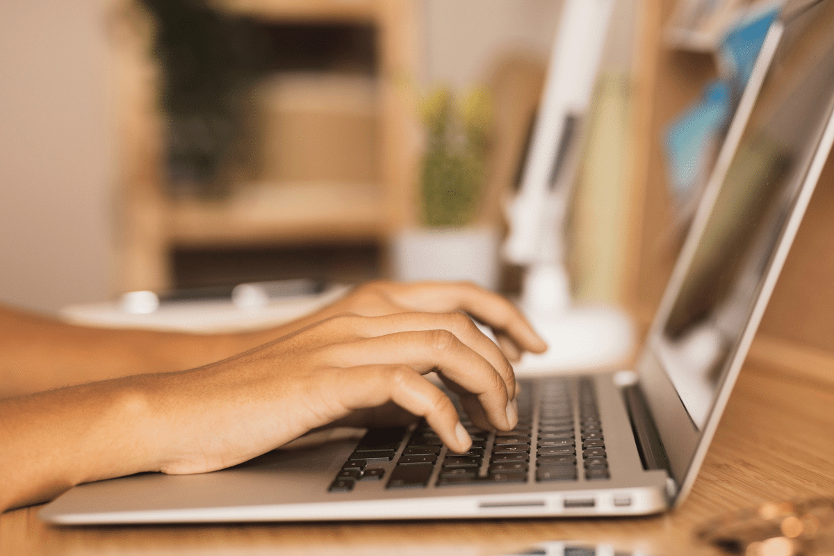 Foto das mãos digitando em teclado de notebook prateado sobre mesa de madeira clara