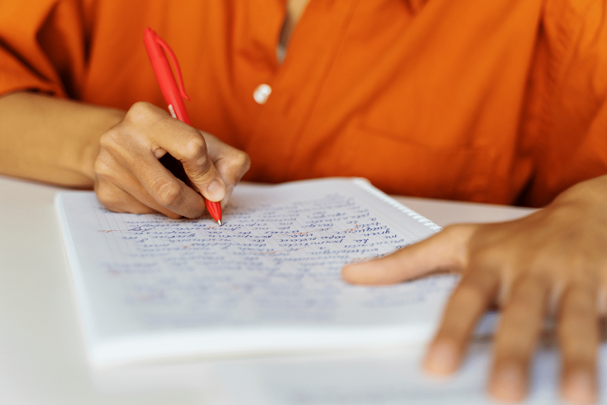 Mão segurando caneta vermelha e escrevendo em caderno com folhas quadriculadas