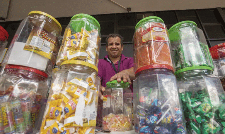 Homem sorridente atrás de potes transparentes cheios de doces embalados à venda em mercado informal.