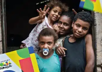 Grupo de crianças sorrindo à porta de casa segurando material do Programa Bolsa Família.