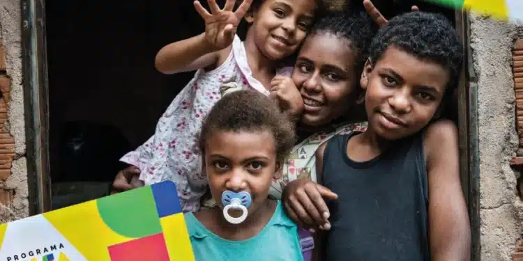 Grupo de crianças sorrindo à porta de casa segurando material do Programa Bolsa Família.