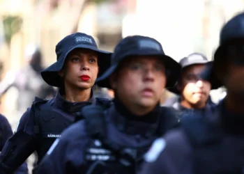 Imagem de policiais militares da Polícia Penal de Minas Gerais em treinamento, com foco em uma mulher com maquiagem e expressão séria, usando uniforme preto e boné característico da corporação.