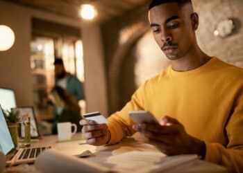 Homem segurando cartão de crédito e celular enquanto observa uma tela de computador em um ambiente de escritório moderno