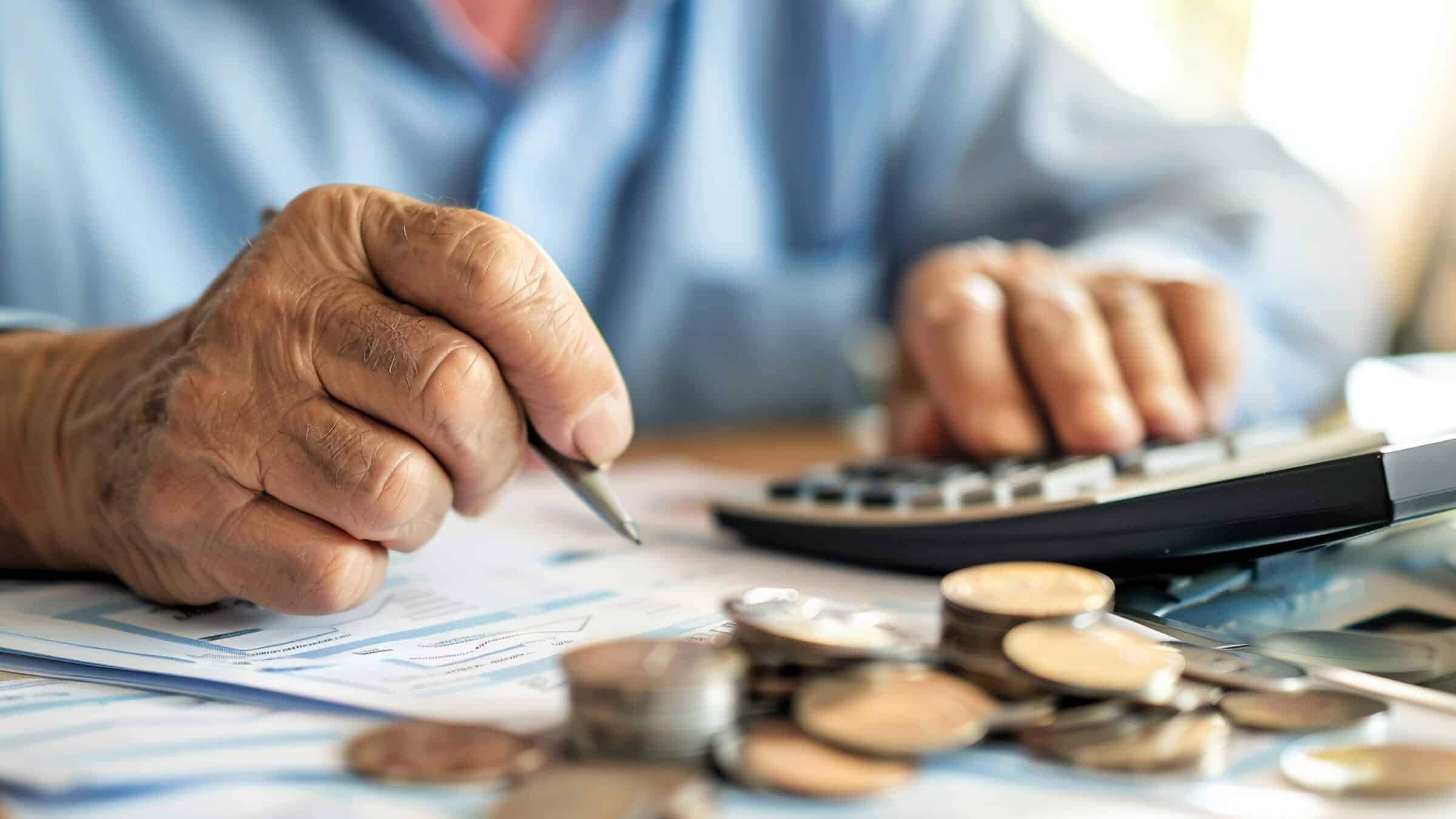 Imagem das mãos de uma pessoa idosa usando calculadora e caneta sobre documentos financeiros, com pilhas de moedas em primeiro plano.