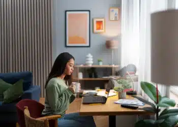 Pessoa concentrada, com uma xícara na mão, estudando em seu escritório, com laptop e materiais de estudo na mesa, ideal para ilustrar a preparação para os concursos de 2026.