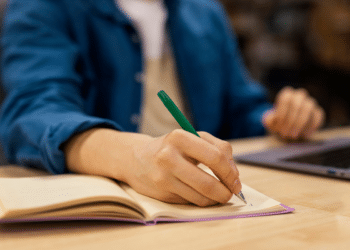 Mão segurando caneta verde escrevendo em caderno aberto sobre mesa com notebook ao fundo