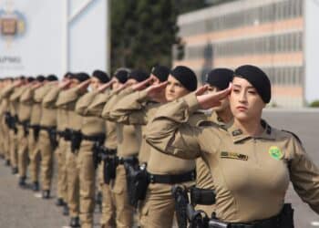 Policiais militares femininas alinhadas em formação realizando saudação com uniforme bege e boina preta