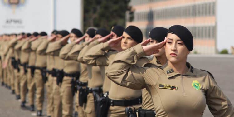 Policiais militares femininas alinhadas em formação realizando saudação com uniforme bege e boina preta
