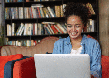 Jovem mulher sorridente usando laptop sentada em sofá vermelho com prateleira de livros ao fundo