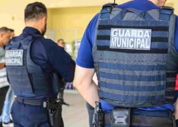 Guardas municipais de costas usando coletes à prova de bala em ambiente interno com outras pessoas ao fundo