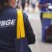 Mulher com colete do IBGE e máscara, carregando bolsa azul com alça amarela em área urbana.