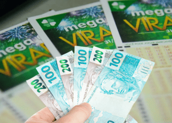 Mão segurando notas de dinheiro com bilhetes de loteria Mega da Virada ao fundo.