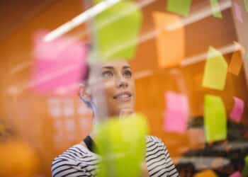 Mulher olhando para post-its rosas, amarelos e verdes em uma parede de vidro em escritório