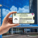 Mão segurando smartphone com carteira de identidade digital na frente do prédio da Previdência Social