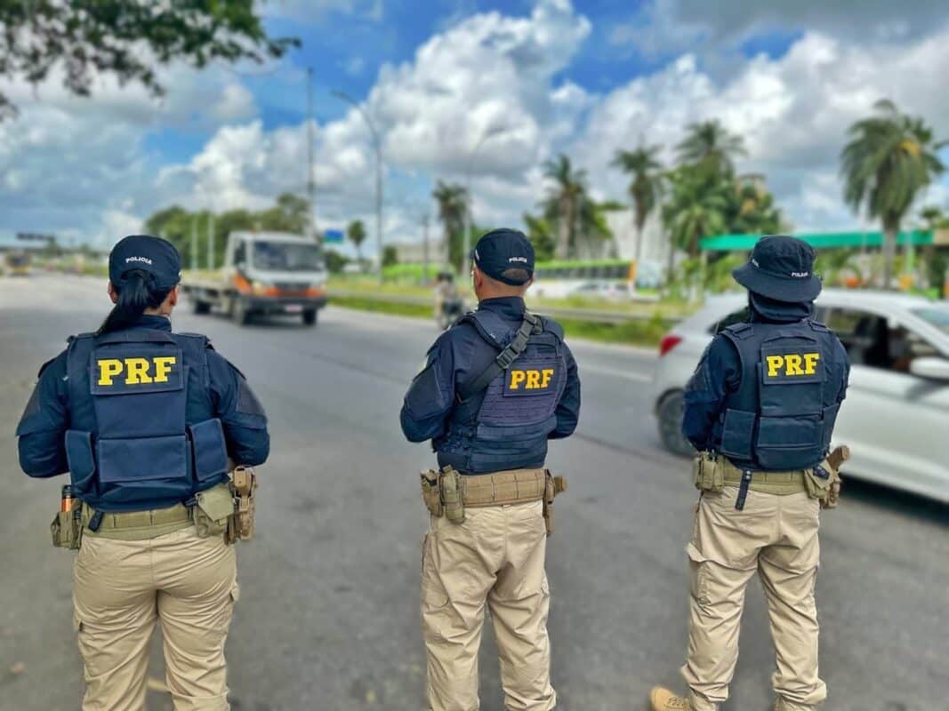 Concurso PRF Três policiais da PRF em operação, fardados com coletes à prova de balas, em rodovia sob céu azul com nuvens