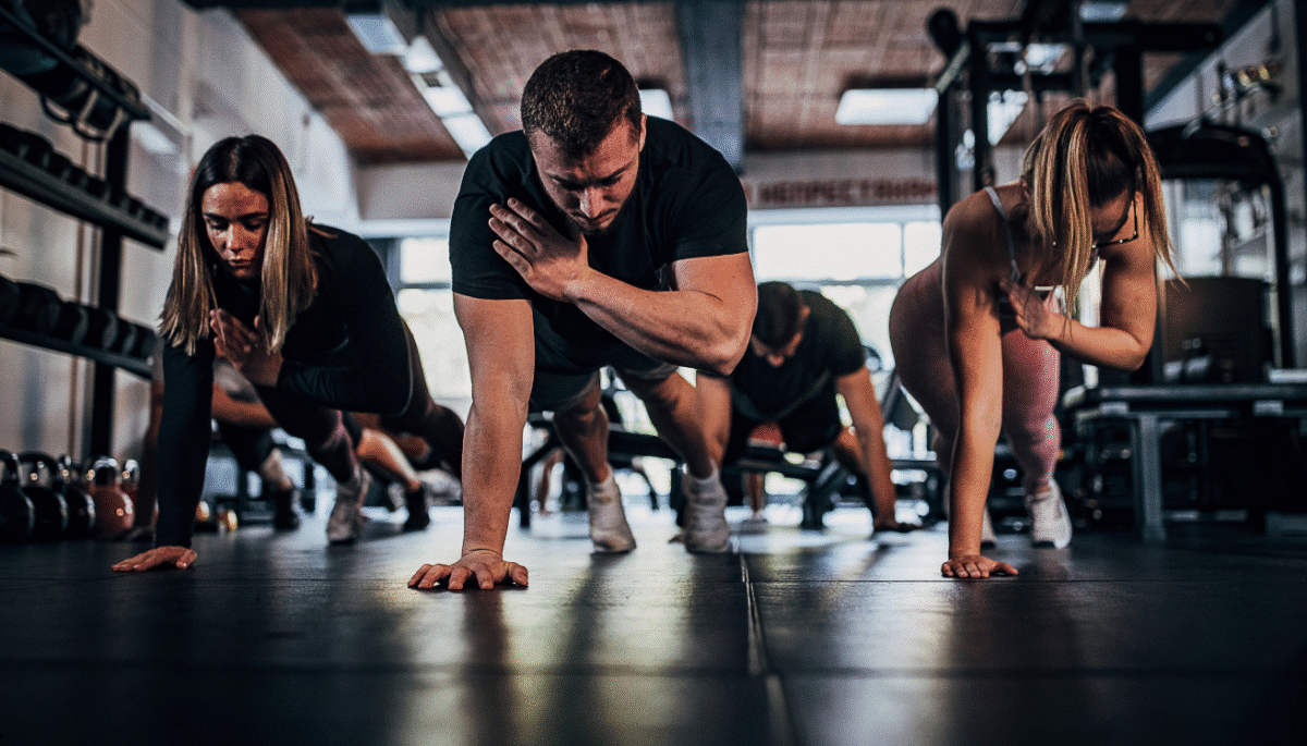 Grupo de pessoas realizando exercício de prancha com um braço na academia, ambiente com equipamentos de musculação.