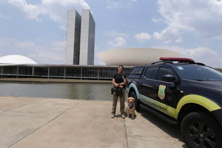 Policial Legislativa Federal ao lado de viatura e cão farejador em frente ao Congresso Nacional em Brasília
