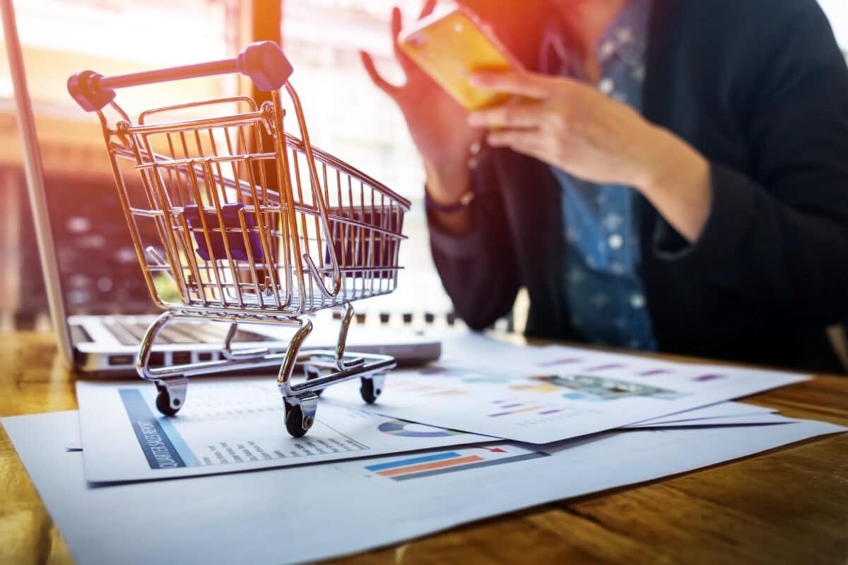 Carrinho de compras em miniatura sobre relatórios financeiros com pessoa usando smartphone ao fundo.