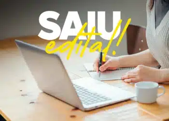 Imagem de uma pessoa escrevendo em um caderno ao lado de um laptop, com destaque para a frase 'SAIU EDITAL!' sobre o concurso da prefeitura, que oferece salários de R$ 9,4 mil.