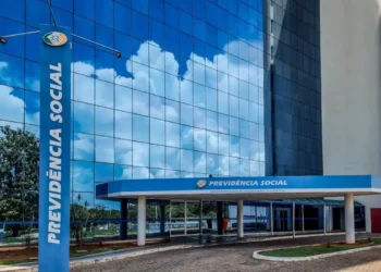 Fachada do prédio do INSS com reflexo do céu e nuvens no vidro, com o letreiro 'Previdência Social' em destaque, indicando o local de atendimento do Instituto Nacional do Seguro Social.