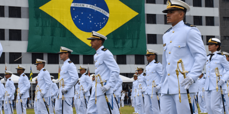 Oficiais da Marinha do Brasil em traje branco durante cerimônia, com grande bandeira do Brasil ao fundo.