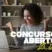 Mulher comemorando notícia positiva em notebook sobre concursos abertos 2026 em ambiente de estudo.
