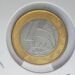 Close-up de uma moeda de 1 real de 2006 segurada por dedos. A imagem destaca a efígie da República no núcleo prateado e o anel dourado com a inscrição "BRASIL" e o ano "2006".