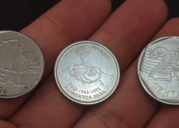 Três moedas brasileiras raras estendidas sobre a palma de uma mão: uma de 1 Real de 1994, uma comemorativa da FAO com o desenho de mãos segurando uma planta e uma de 25 centavos de 1995.