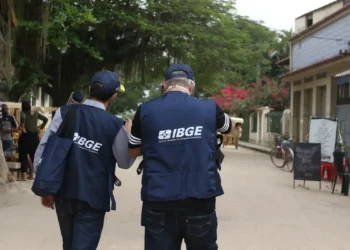 Dois recenseadores do IBGE com coletes azuis caminhando em rua com árvores, lojas e bicicletas