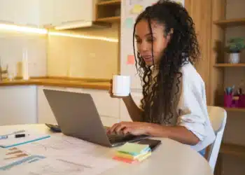 Profissional trabalhando de casa, analisando gráficos e tomando café em um ambiente doméstico com laptop.
