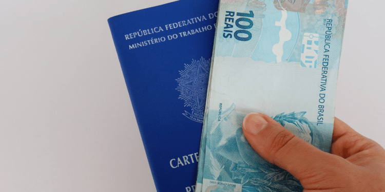 Mão segurando uma nota de 100 reais em frente a uma carteira de trabalho azul do Brasil
