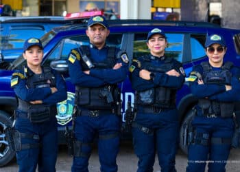 Quatro guardas municipais uniformizados posam com braços cruzados diante de viatura azul