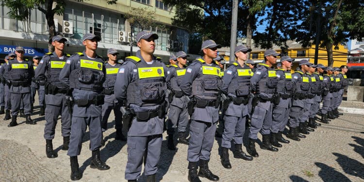 Grupo de policiais militares em uniforme cinza alinhados em praça durante solenidade