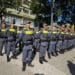Grupo de policiais militares em uniforme cinza alinhados em praça durante solenidade