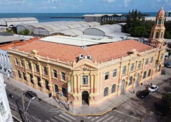 Edifício antigo com fachada amarela e rosa em esquina, próximo ao mar azul e céu parcialmente nublado