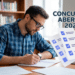 Homem escrevendo em caderno sobre mesa com folhas e livros, ao lado de ilustração de provas e caneta