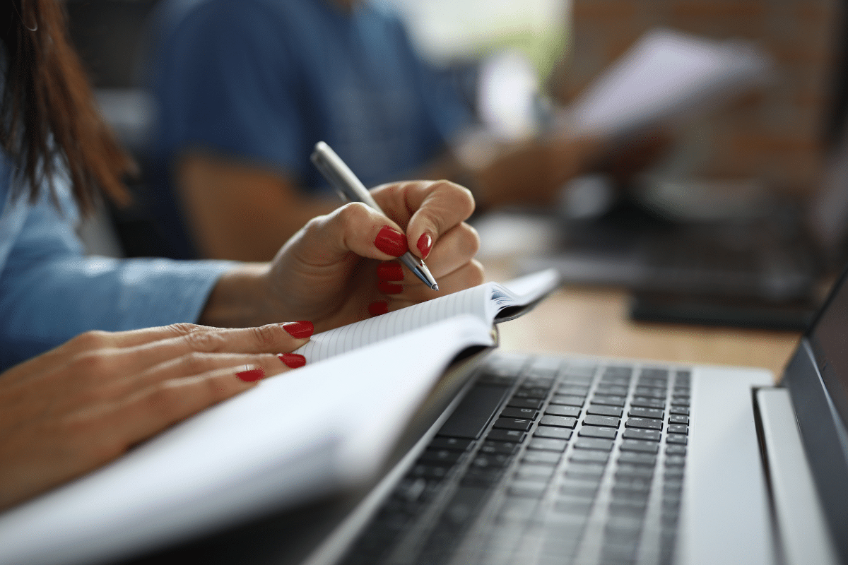 Mulher escreve em caderno aberto sobre notebook enquanto estuda