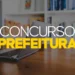 Notebook sobre mesa de madeira com o texto "Concurso Prefeitura" em destaque sobre fundo escurecido com estante ao fundo