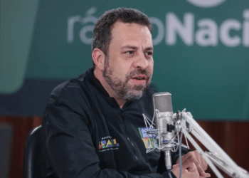 Homem de barba fala ao microfone profissional em estúdio, com jaqueta do Governo do Brasil