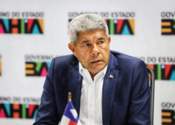Homem de cabelos grisalhos com terno azul sentado diante de painel do Governo da Bahia