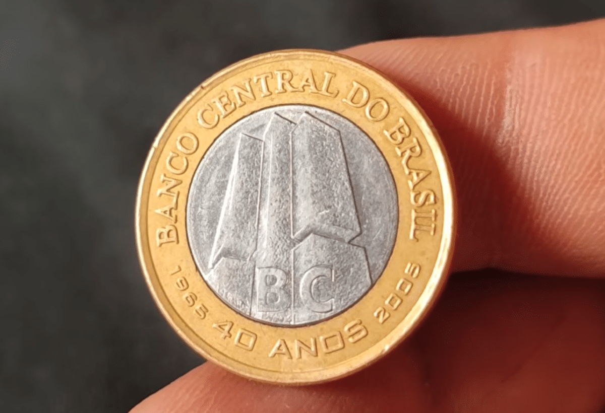 Visão frontal detalhada da moeda de 1 real comemorativa dos 40 anos do Banco Central, segurada entre os dedos. No centro prateado, vê-se o edifício-sede do BC e a sigla "BC". No anel dourado, a inscrição "BANCO CENTRAL DO BRASIL" e "1965 40 ANOS 2005".