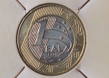 Fotografia em macro de uma moeda de 1 real brasileira do ano de 1998, centralizada em um suporte de papel branco (coin holder). A imagem destaca o número "1", a palavra "REAL" e o ano "1998" gravados no núcleo prateado, rodeado pelo anel dourado com grafismos indígenas marajoaras e estrelas do Cruzeiro do Sul.