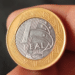 Close-up do anverso de uma moeda de 1 real comemorativa dos 40 anos do Banco Central (2005). O centro prateado exibe o prédio do Banco Central e a sigla BC, enquanto o anel dourado contém a legenda "BANCO CENTRAL DO BRASIL" e as datas "1965 40 ANOS 2005".