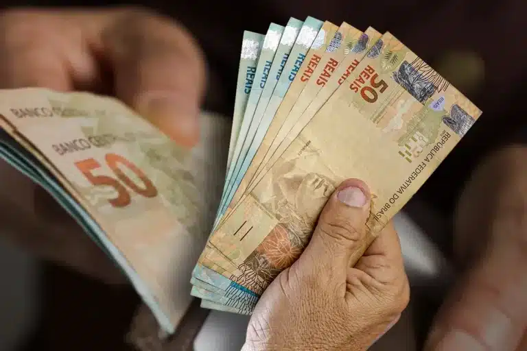 Notas de 50 reais sendo seguradas, representando o pagamento do BPC de fevereiro, com a dúvida sobre a possível liberação do 13º salário.