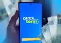 Imagem do aplicativo Caixa Tem sendo acessado em um smartphone, com dinheiro em notas ao fundo. O aplicativo vai liberar R$ 1.200 para diversos brasileiros até 5 de março.